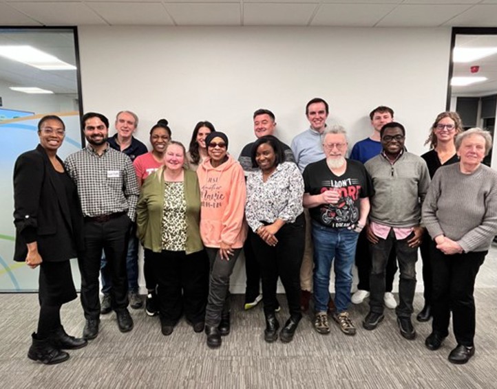 Group photo of the Resident Forum at NHG’s office. In the front row, seated left to right: Emma-Louise (resident Board member and Chair of the Resident Forum), Kautuk, Susan, Lin, Lacey, David, Ebenezer, and Mary. In the back row, standing left to right: Ian, Chioma, Rebecca, Robert, Gareth, Conrad, and Symmie (resident Board member). The group is smiling and facing the camera in a bright, modern meeting space, reflecting a collaborative and resident-led environment